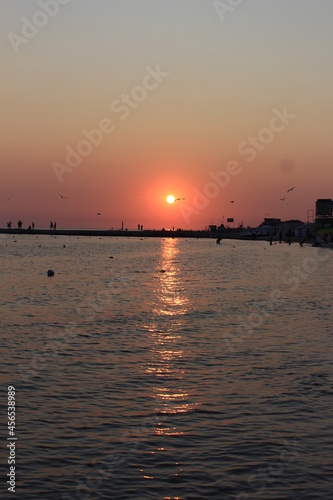 Sunset paints the sky and sea in shades of red. Seagulls fly against the backdrop of the setting sky and the setting sun. The sea pier is visible in the distance. The resting people watch the sunset © Yuliya