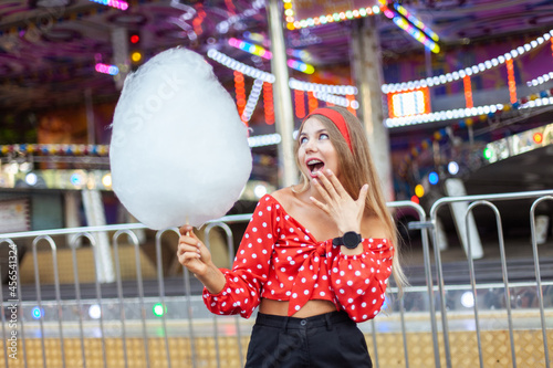 Adorable cute beautiful woman with cotton candy stands in the middle of an amusement park with bright colors, positive and cheerful, happy and optimistic emotion