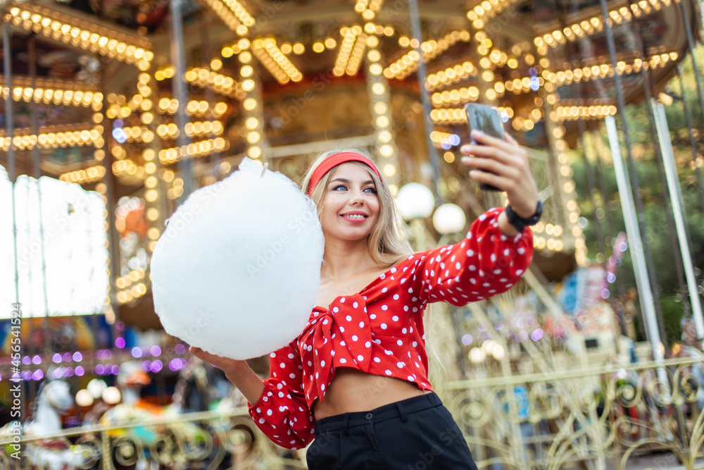 Adorable cute beautiful woman with cotton candy doing selfie on phone while stands in the middle of an amusement park with bright colors, positive and cheerful, happy and optimistic emotion