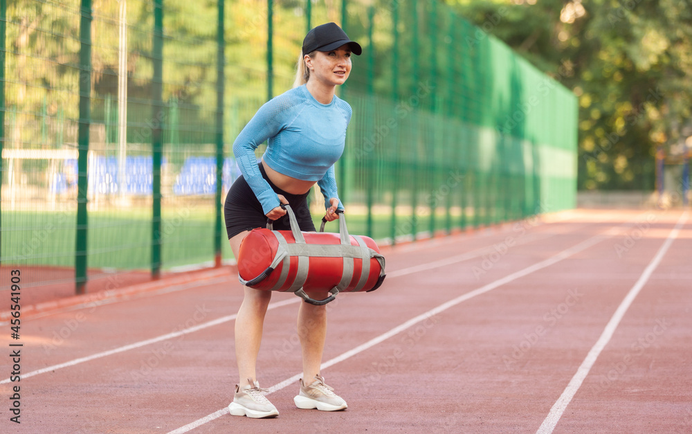Young athletic woman workout with a heavy sand bag at the stadium in the morning