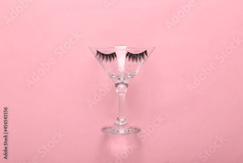 Minimal party layout. Cocktail glass with false eyelashes on a pink pastel background. Top view