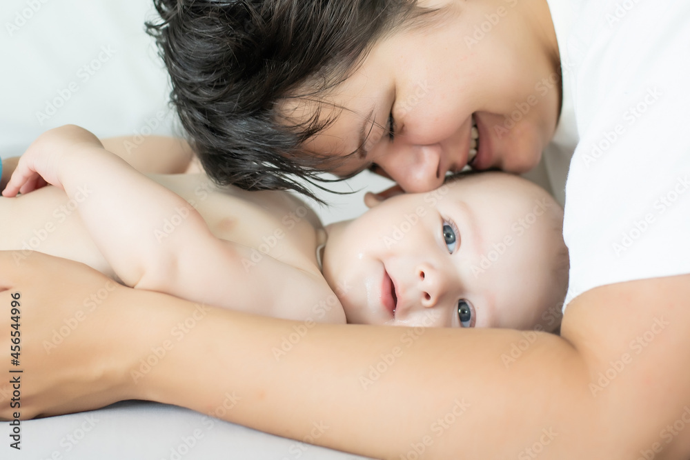 Mother and child on a white bed. Mom and baby boy in diaper playing in bedroom. Parent and little kid relaxing at home. Family having fun together.