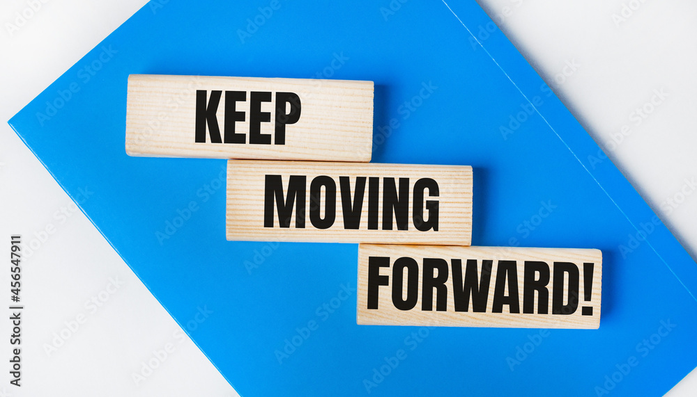 There is a blue notebook on a light gray background. Above are three wooden blocks with the words KEEP MOVING FORWARD There is a blue notebook on a light gray background. Above are three wooden blocks with the words KEEP MOVING FORWARD