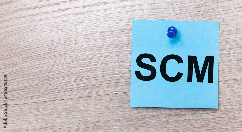 On a light wooden background - a light blue square sticker with the text SCM Supply Chain Management On a light wooden background - a light blue square sticker with the text SCM Supply Chain Management