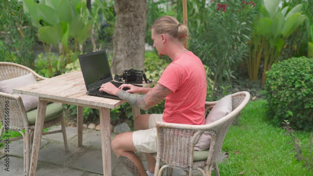 Man photogrpher in pink shirt working in outdoor cafe on laptop. Handheld shot