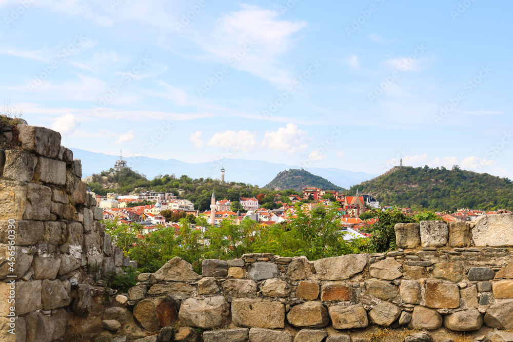 Stone fortress of the ancient city of Plovdiv and panoramic view of the landscape with the buildings and trees, the hills, the mountains in the background, and a blue sky - medieval tourist postcard