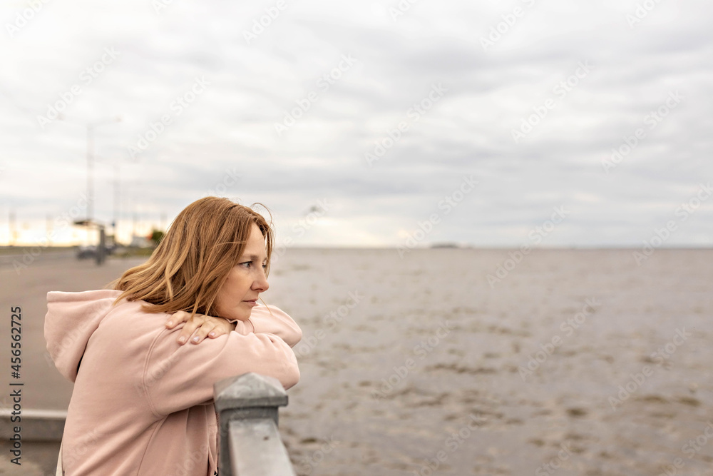 A young woman on a cloudy evening stands on the embankment and looks at the sea