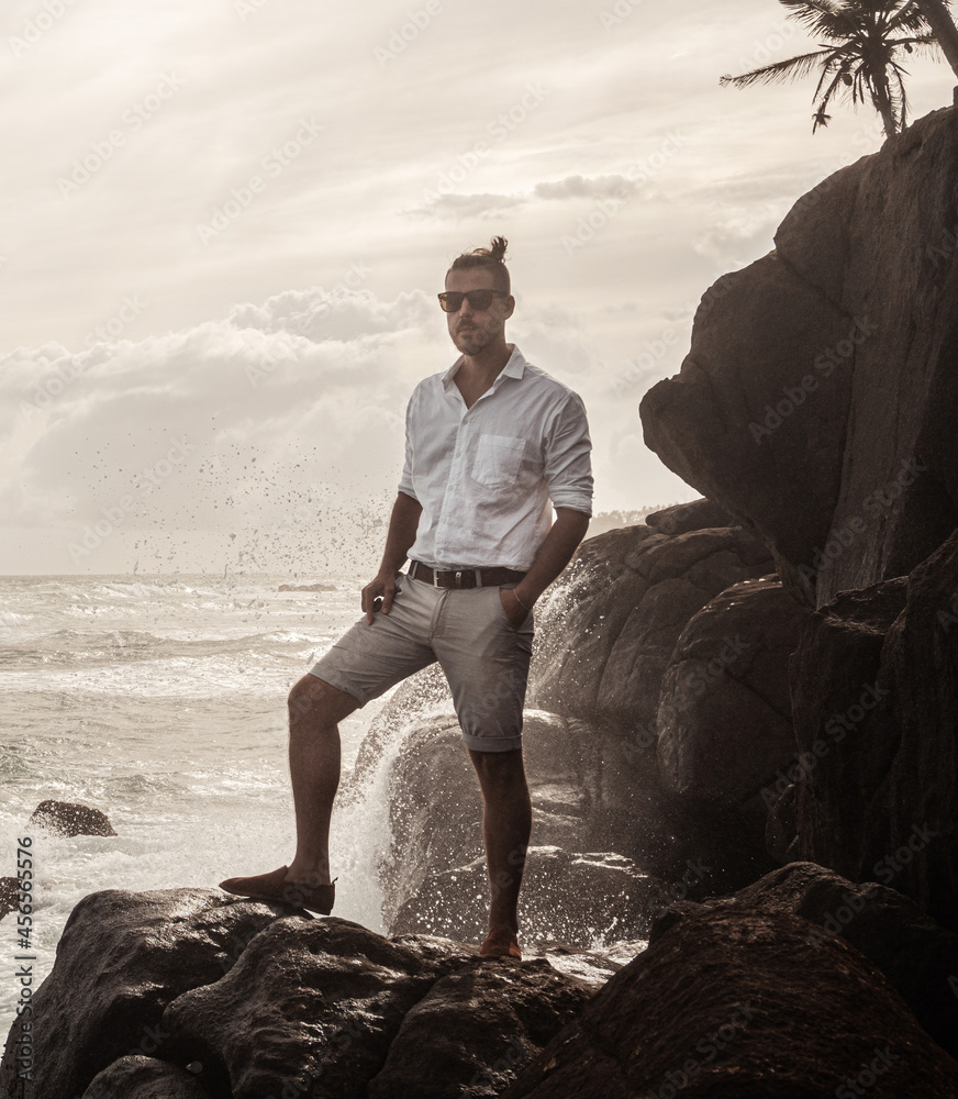 Mężczyzna elegancko ubrany stojący na skałach na tle oceanu i wzburzonych fal. - obrazy, fototapety, plakaty 