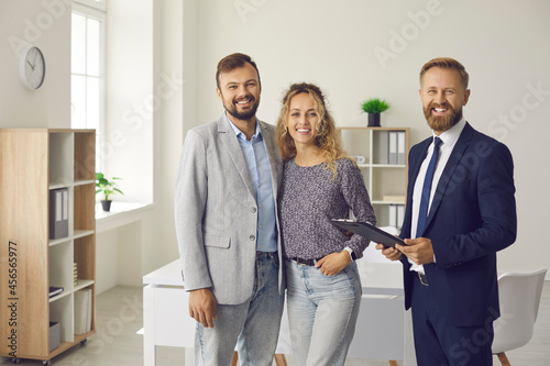 Portrait of happy clients and real estate agent. Young married couple together with realtor, bank specialist or mortgage broker standing in office, smiling and looking at camera photo