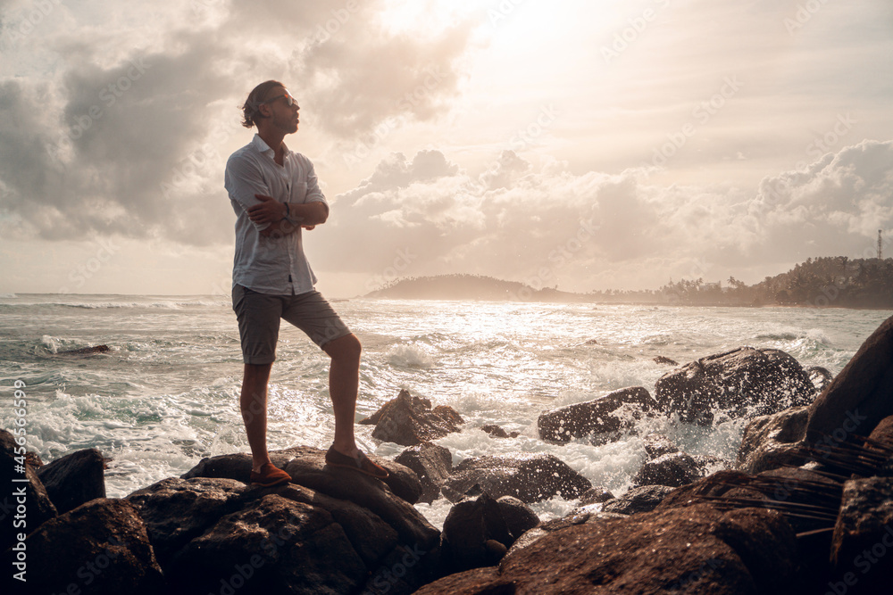 Mężczyzna elegancko ubrany stojący na skałach na tle oceanu i wzburzonych fal. - obrazy, fototapety, plakaty 