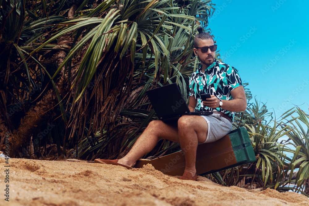 Cyfrowy nomada, mężczyzna pracujący na tropikalnej plaży z laptopem i telefonem. - obrazy, fototapety, plakaty 