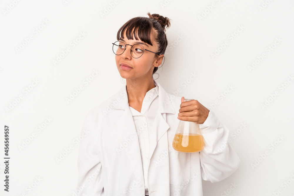 Young mixed race scientist woman holding a test tube isolated on white background  looks aside smiling, cheerful and pleasant.