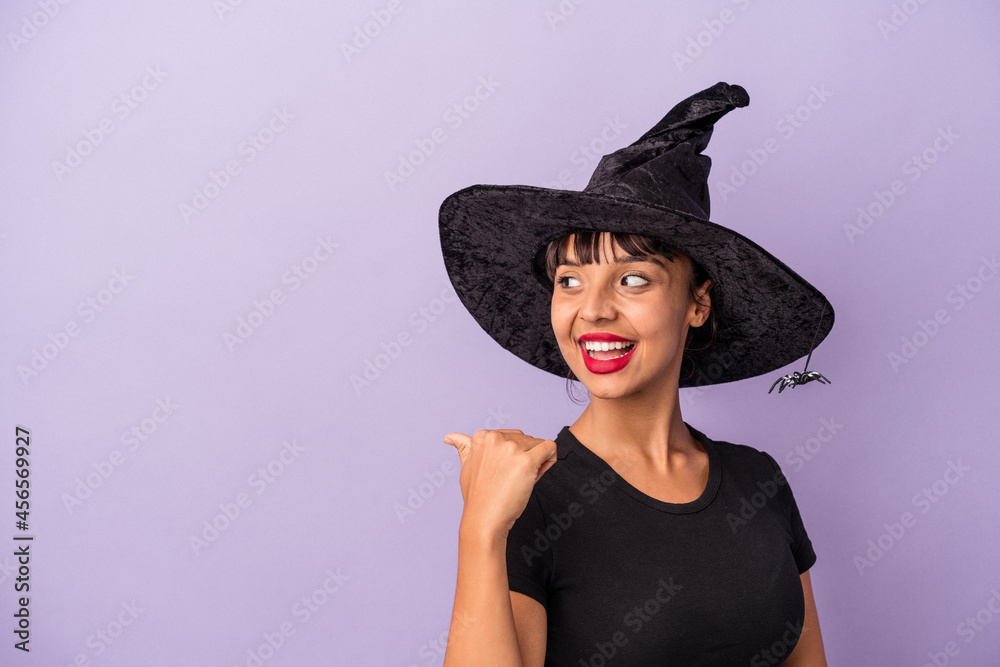 Young mixed race woman disguised as a witch isolated on purple background points with thumb finger away, laughing and carefree. Young mixed race woman disguised as a witch isolated on purple background points with thumb finger away, laughing and carefree.