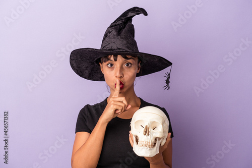 Bild auf Leinwand Young mixed race woman disguised as a witch holding a skull isolated on purple background  keeping a secret or asking for silence