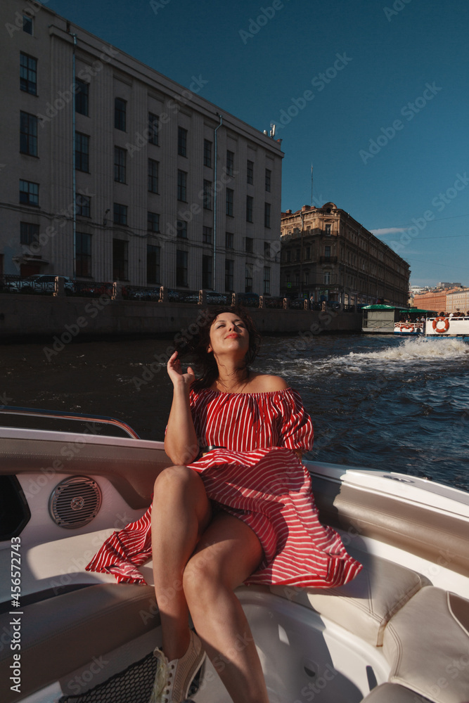 brunette woman in developing red striped dress sits happy in boat sailing along canal in St. Petersburg, Russia