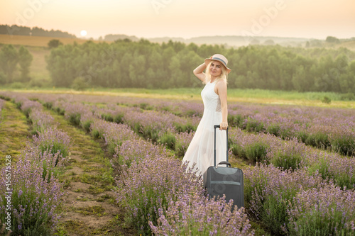 Full length portrait of pretty young lady, wearing light dress, straw hat, walking with bag in summer flowering lavender field, enjoying scent of lavender