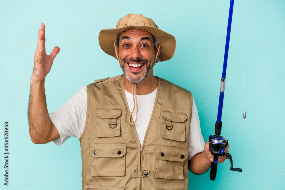 Middle age caucasian fisherman holding rod isolated on blue background receiving a pleasant surprise, excited and raising hands. Middle age caucasian fisherman holding rod isolated on blue background receiving a pleasant surprise, excited and raising hands.