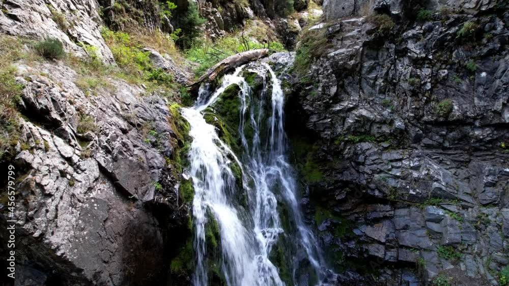 A mountain waterfall in a rocky gorge. The view from the drone is of steep cliffs, moss, green plants and the river. There are stones lying. Splashes from the water. There is a log lying. Almaty A mountain waterfall in a rocky gorge. The view from the drone is of steep cliffs, moss, green plants and the river. There are stones lying. Splashes from the water. There is a log lying. Almaty