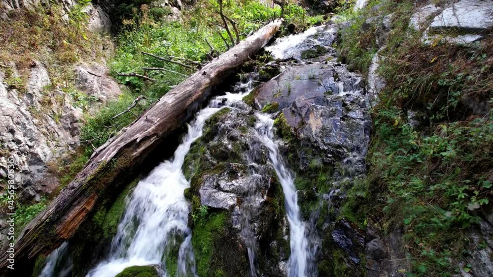 A mountain waterfall in a rocky gorge. The view from the drone is of steep cliffs, moss, green plants and the river. There are stones lying. Splashes from the water. There is a log lying. Almaty A mountain waterfall in a rocky gorge. The view from the drone is of steep cliffs, moss, green plants and the river. There are stones lying. Splashes from the water. There is a log lying. Almaty