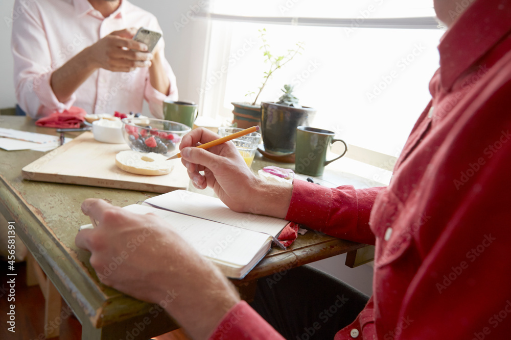 Fototapeta premium Cropped view of men sitting at dining table drinking orange juice looking at smartphone, writing in notebook