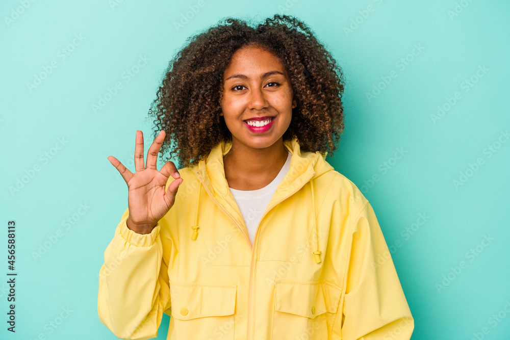 Young african american woman with curly hair isolated on blue background cheerful and confident showing ok gesture. Young african american woman with curly hair isolated on blue background cheerful and confident showing ok gesture.