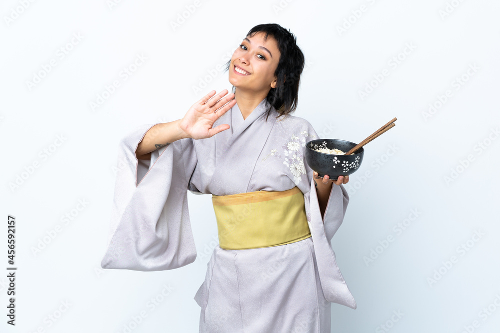 Young woman wearing kimono holding a bowl of noodles over isolated white background saluting with hand with happy expression Young woman wearing kimono holding a bowl of noodles over isolated white background saluting with hand with happy expression