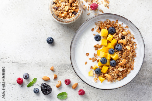 Granola (muesli) and yogurt in bowl, nuts, fresh berries blueberry and mango fruit. Healthy eating. Easy breakfast or snack. Top view, copy space, light grey background.