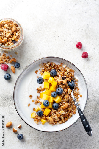 Granola (muesli) and yogurt in bowl, nuts, fresh berries blueberry and mango fruit. Healthy eating. Easy breakfast or snack. Top view, copy space, light grey background.