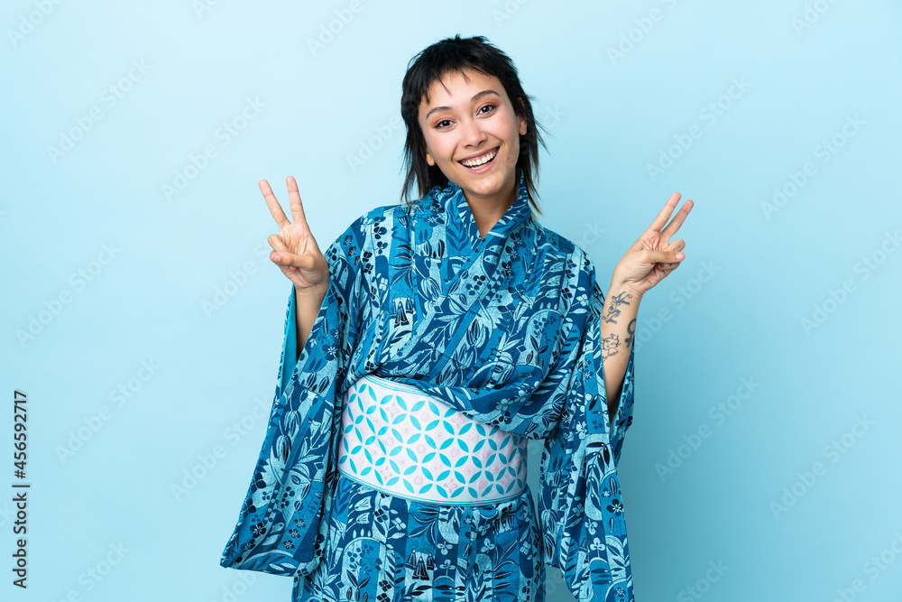 Young woman wearing kimono over isolated blue background showing victory sign with both hands Young woman wearing kimono over isolated blue background showing victory sign with both hands