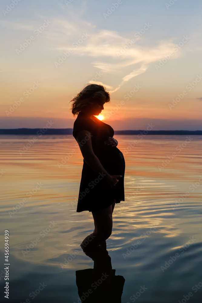 Silhouette of a female expecting a baby, who stands in the water of the lake and touch her belly against the background of sunset.