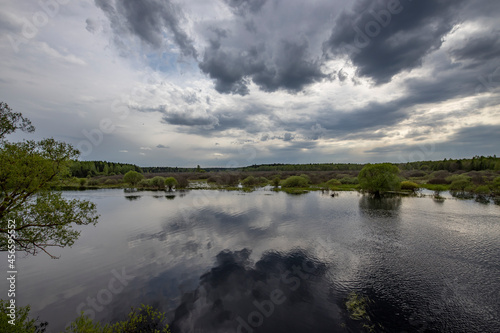 Landscape with a dramatic sky reflected in the river. Early spring  juicy May greens. Bright green foliage on trees and bushes.