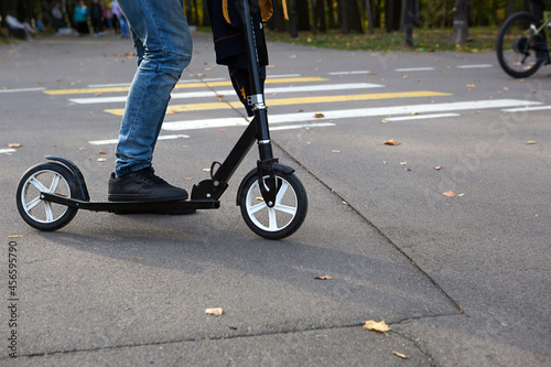 The legs of a man in jeans and sneakers on a scooter in the park in autumn with fallen dry yellow leaves on the asphalt. Autumn walks, active lifestyle, eco-friendly transport, traffic