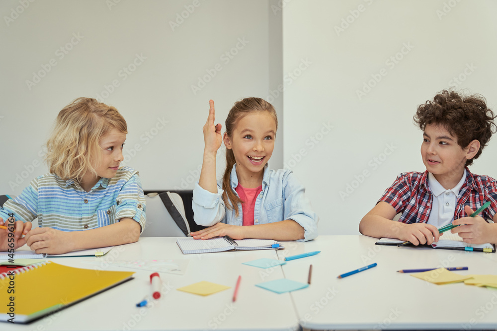 Smart little schoolgirl looking away raised her hand while studying, sitting at the table in elementary school classroom with other kids Smart little schoolgirl looking away raised her hand while studying, sitting at the table in elementary school classroom with other kids