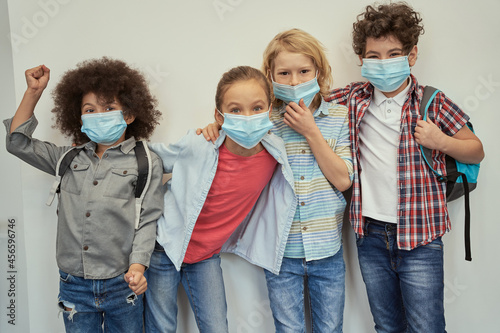 Enthusiastic pupils. Charming diverse kids wearing protective face masks looking at camera, posing together over light background