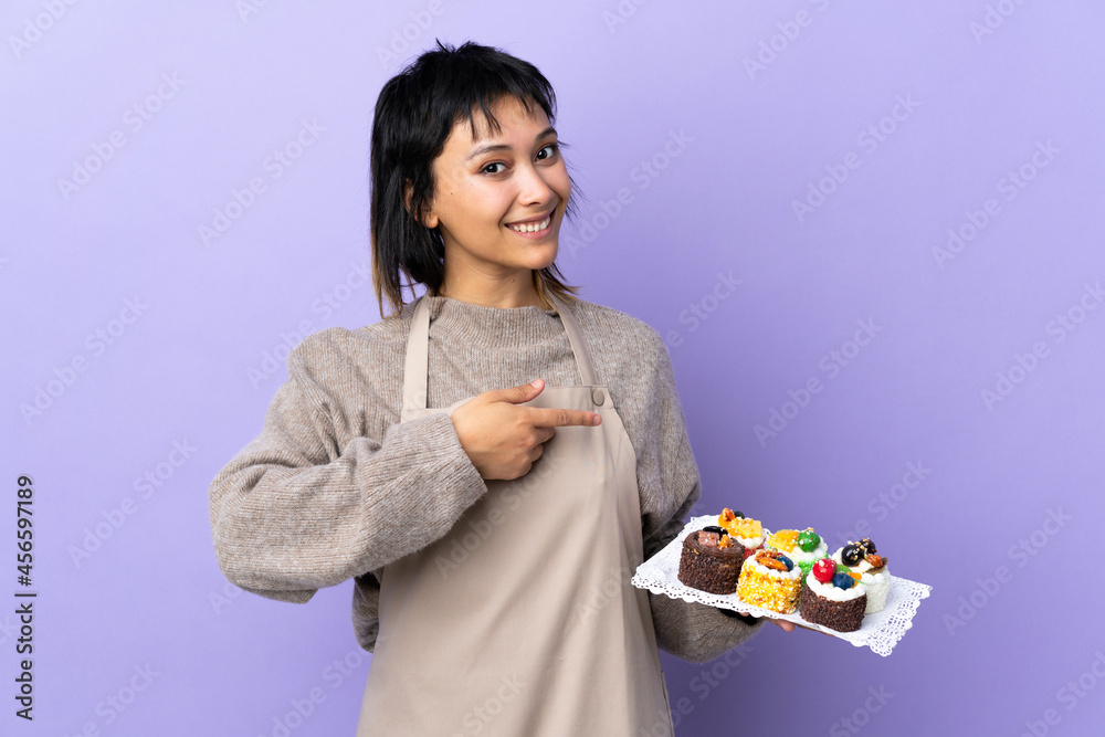 Young Uruguayan woman holding lots of different mini cakes over isolated purple background pointing finger to the side Young Uruguayan woman holding lots of different mini cakes over isolated purple background pointing finger to the side