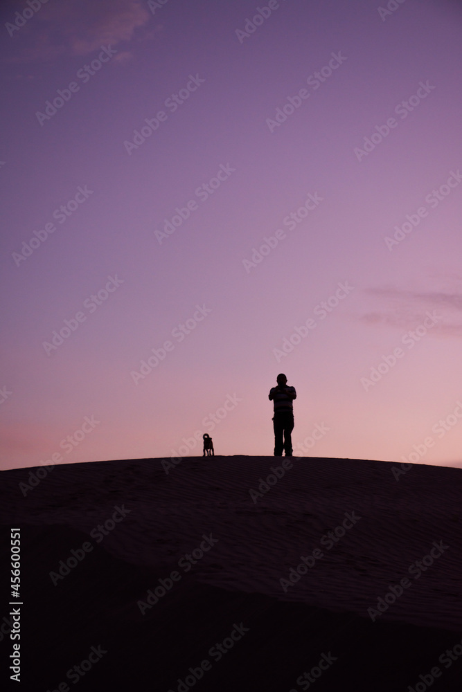 Silhouette of a man and his dog on the top of a dune at sunset
