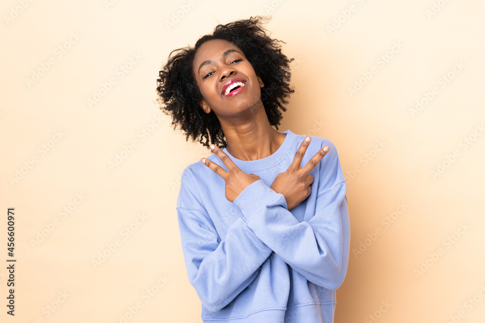 Fototapeta premium Young African American woman isolated on beige background smiling and showing victory sign