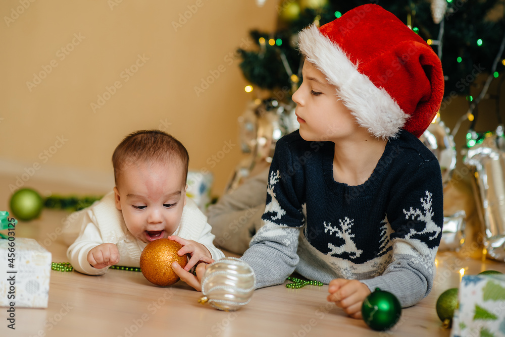 Fototapeta premium A little boy and his brother play with gifts under a festive Christmas tree. Happy Christmas and New Year celebrations