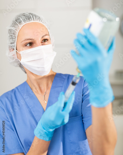 Masked female nurse working at the hospital fills a syringe with saline for injection in the treatment room. Close-up ..portrait