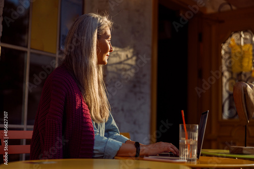 Positive mature Asian lady with long hair types on contemporary laptop at small round table on outdoors cafe terrace at sunset side view