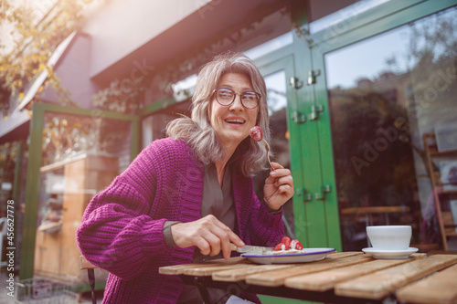 Smiling grey haired senior woman guest in purple jacket eats tasty strawberry dessert sitting at table on outdoors cafe terrace on autumn day