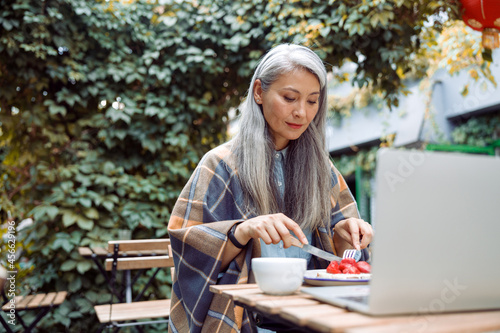 Positive mature Asian woman eats delicious dessert with fresh strawberries in front of open laptop at table on outdoors cafe terrace on autumn day