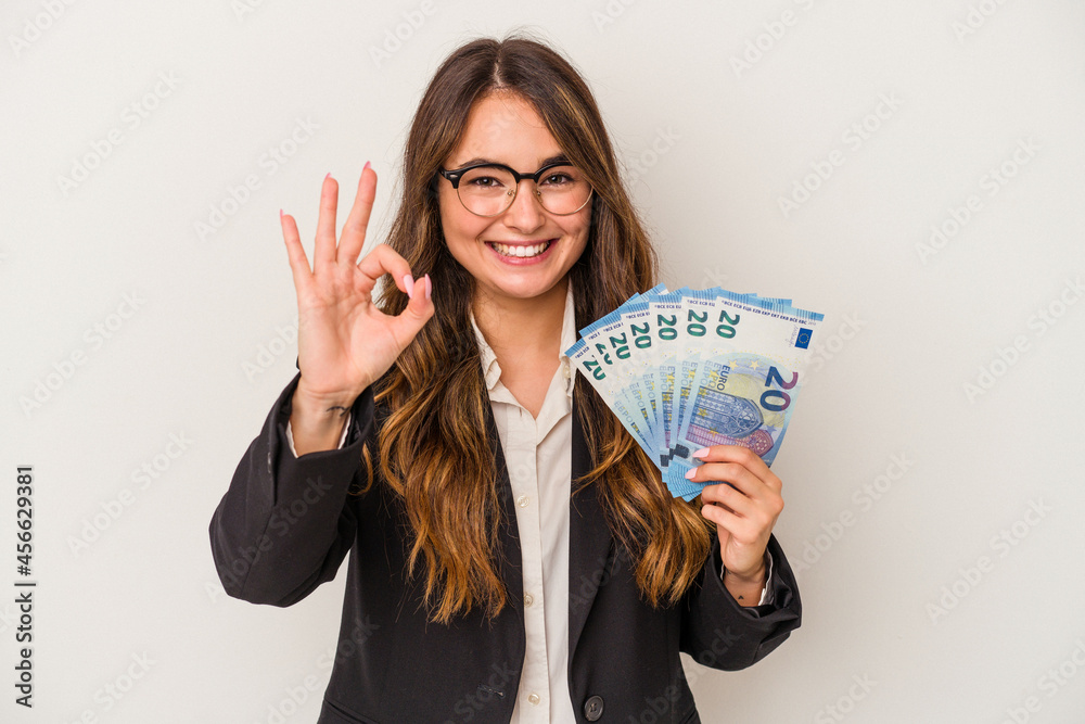 Young caucasian business woman holding banknotes isolated on white background cheerful and confident showing ok gesture. Young caucasian business woman holding banknotes isolated on white background cheerful and confident showing ok gesture.