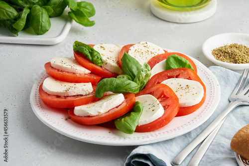 Appetizing italian caprese salad with sliced tomatoes, mozzarella, basil, olive oil, pepper and salt on white plate. Mediterranean healthy fresh appetizer. Horizontal side view of rustic meal