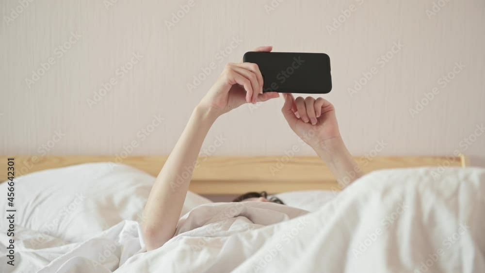 Young woman under warm blanket watches video via cellphone lying in large bed in light room close view. Internet addiction disease