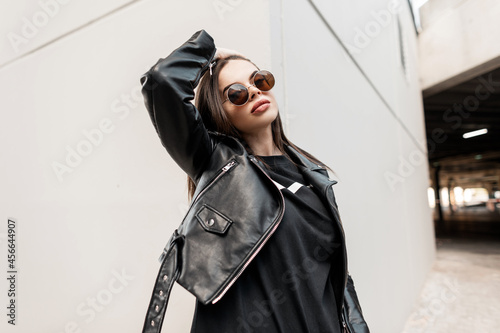 Urban beautiful young hipster woman with stylish round sunglasses in a fashion leather jacket with a black sweatshirt walks in the city