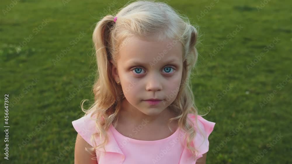Little girl in a pink dress close up. She stands on the green grass in the park and smiles sincerely. She has blonde wavy hair and honest blue eyes Little girl in a pink dress close up. She stands on the green grass in the park and smiles sincerely. She has blonde wavy hair and honest blue eyes
