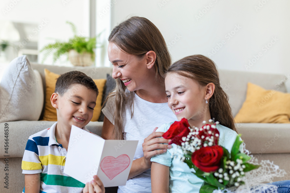Young mother with a bouquet of roses laughs, hugging her son, and сheerful girl with a card and roses congratulates mom during holiday celebration at home. mothers day