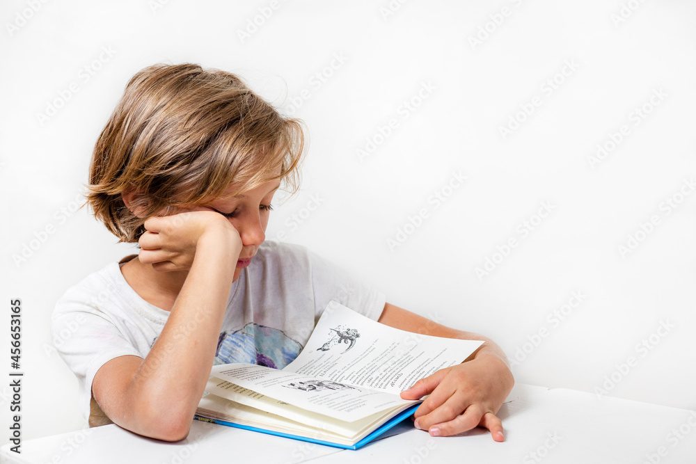 serious boy reading an interesting book at a desk against a background of a white wall, back to school, Authenticity serious boy reading an interesting book at a desk against a background of a white wall, back to school, Authenticity