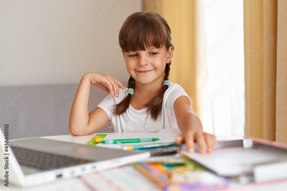 Portrait of happy smiling schoolchild, wearing white t shirt sitting at table against window with curtains in front of laptop, having positive expression, doing homework. Portrait of happy smiling schoolchild, wearing white t shirt sitting at table against window with curtains in front of laptop, having positive expression, doing homework.